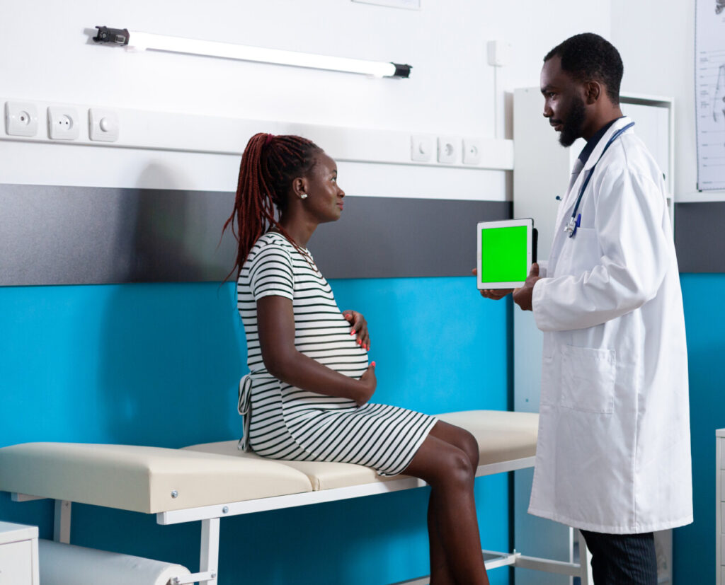 Physician showing tablet with vertical green screen to pregnant woman. Adult expecting baby looking at chroma key with isolated template and mockup background for healthcare.